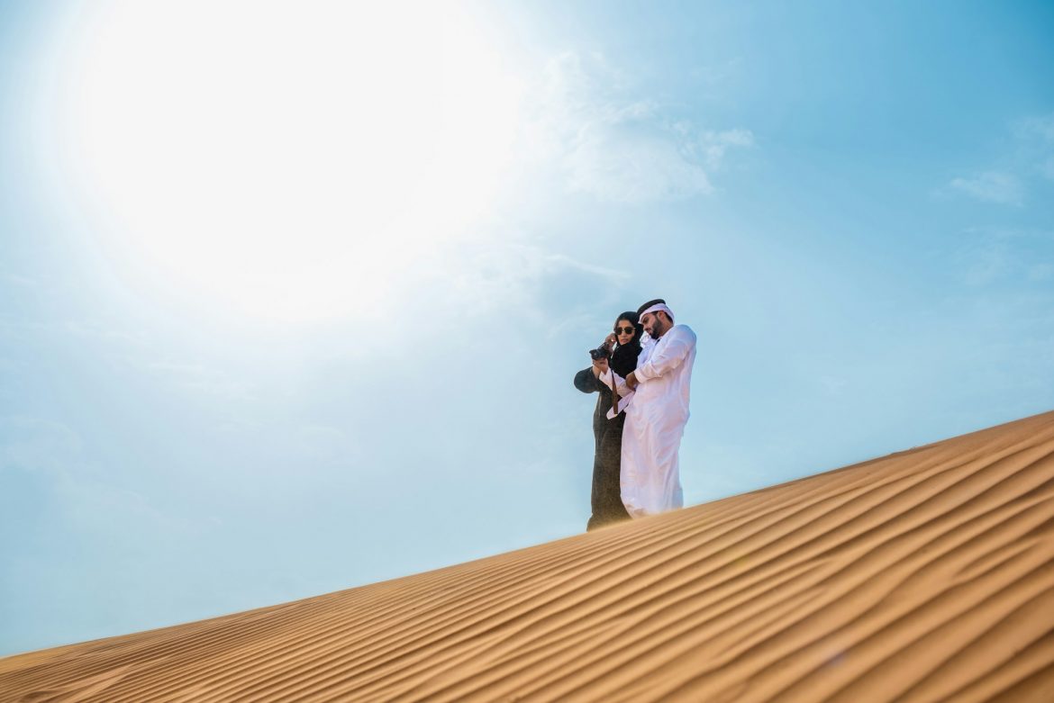 Middle eastern couple wearing traditional clothes taking smartphone selfie on desert dune, Dubai,